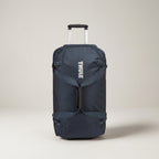 A navy blue Thule rolling duffel bag with an extended silver telescoping handle, shown against a neutral light beige studio background.
