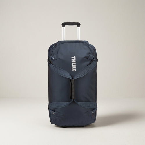 A navy blue Thule rolling duffel bag with an extended silver telescoping handle, shown against a neutral light beige studio background.