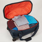 An open navy blue rolling suitcase with a bright orange interior, shown packed with folded clothing including a light gray button-down shirt, a bright blue shirt, a red-and-white checkered shirt, and a dark maroon item. The suitcase is photographed on a clean light gray studio background.