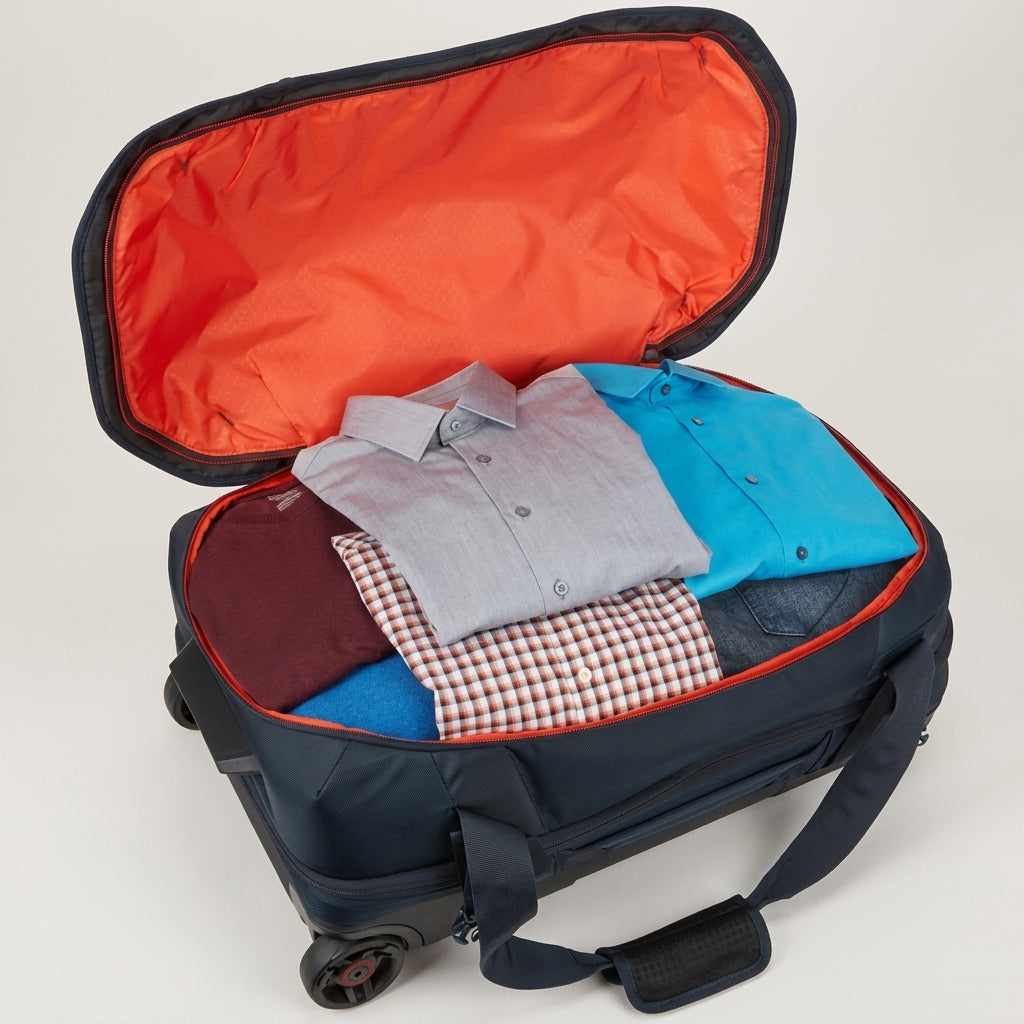 An open navy blue rolling suitcase with a bright orange interior, shown packed with folded clothing including a light gray button-down shirt, a bright blue shirt, a red-and-white checkered shirt, and a dark maroon item. The suitcase is photographed on a clean light gray studio background.