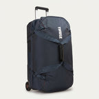 A navy blue Thule rolling duffel bag with an extended telescoping handle and wheels, shown from a three-quarter angle against a light off-white background.