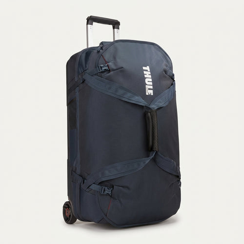 A navy blue Thule rolling duffel bag with an extended telescoping handle and wheels, shown from a three-quarter angle against a light off-white background.